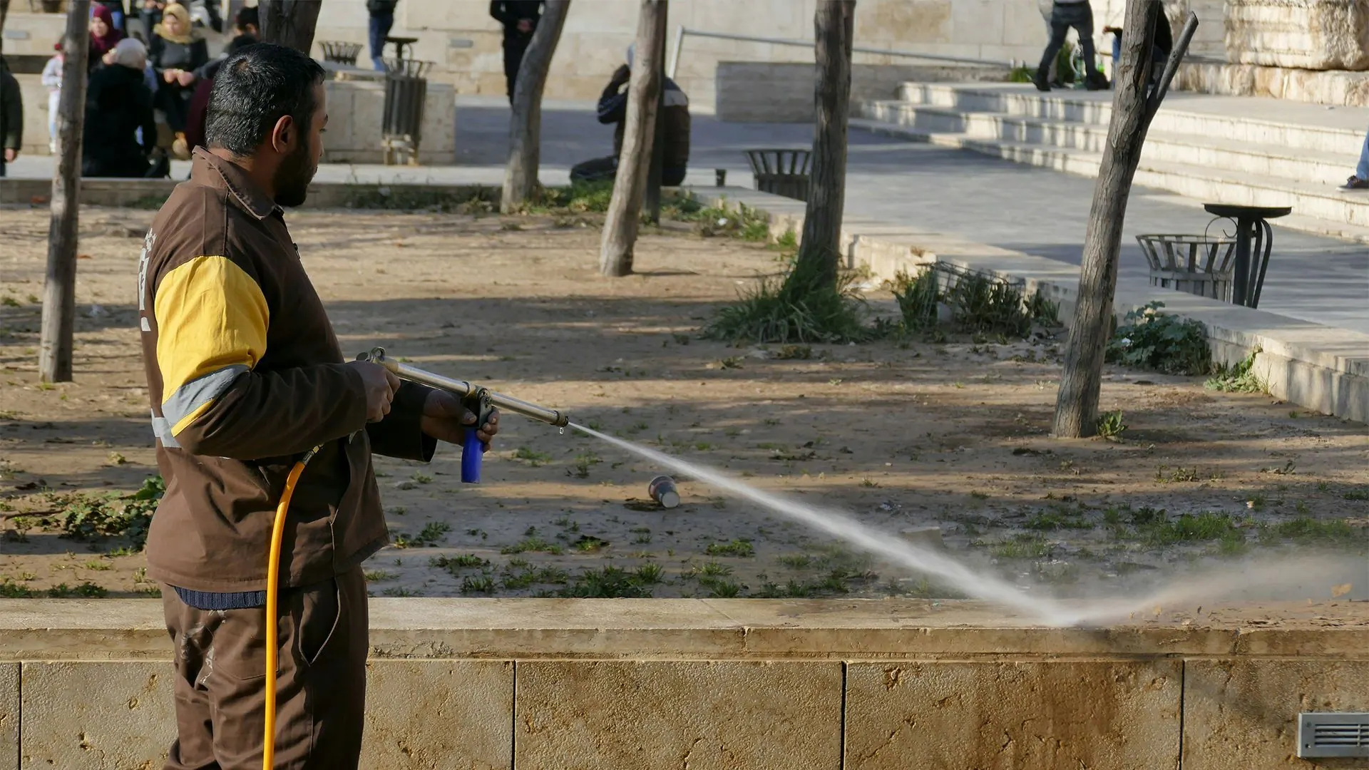 Person spraying water with a hose along a sidewalk in a park area.