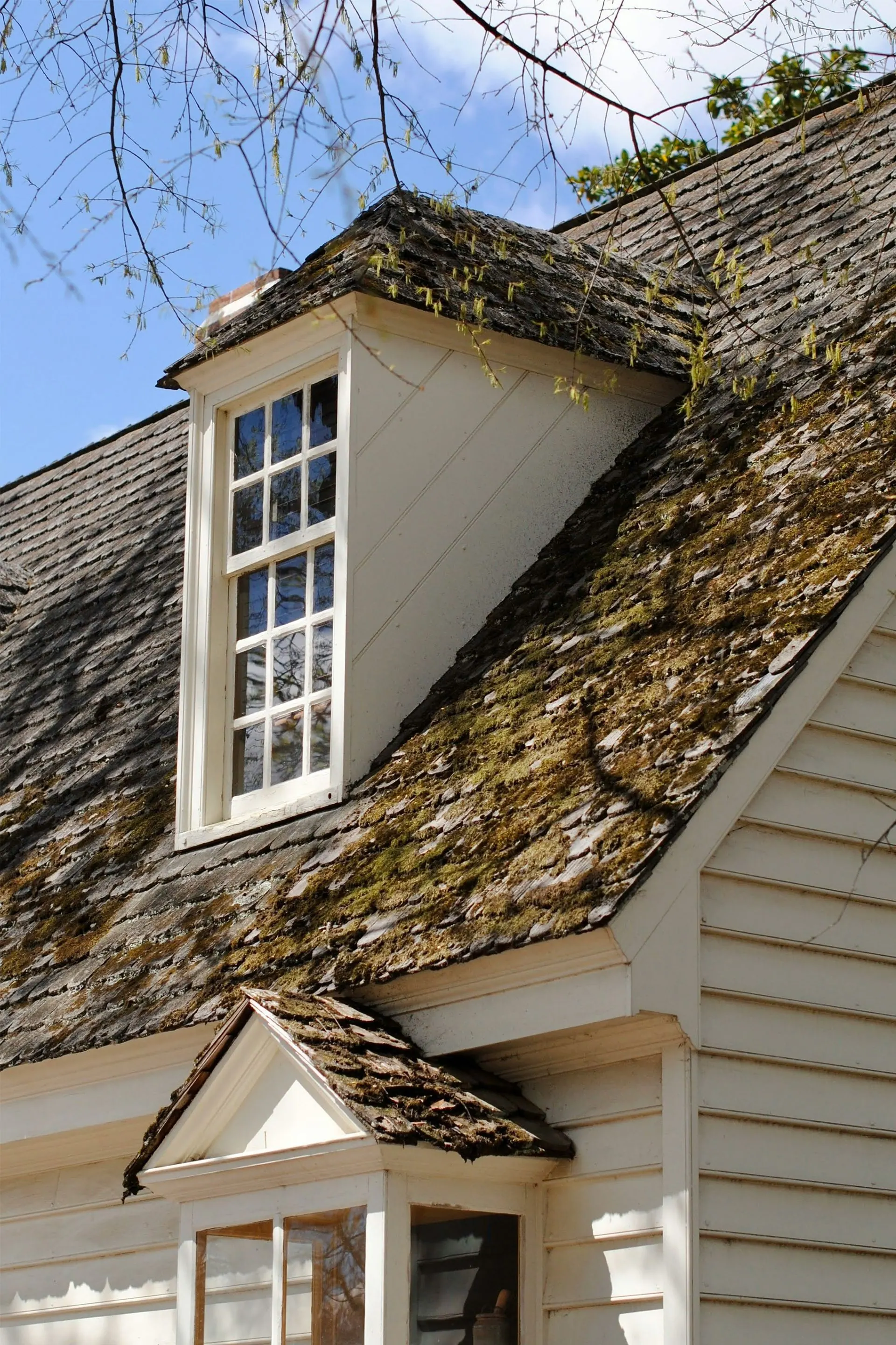 White dormer window on a weathered shingled house roof under a blue sky.